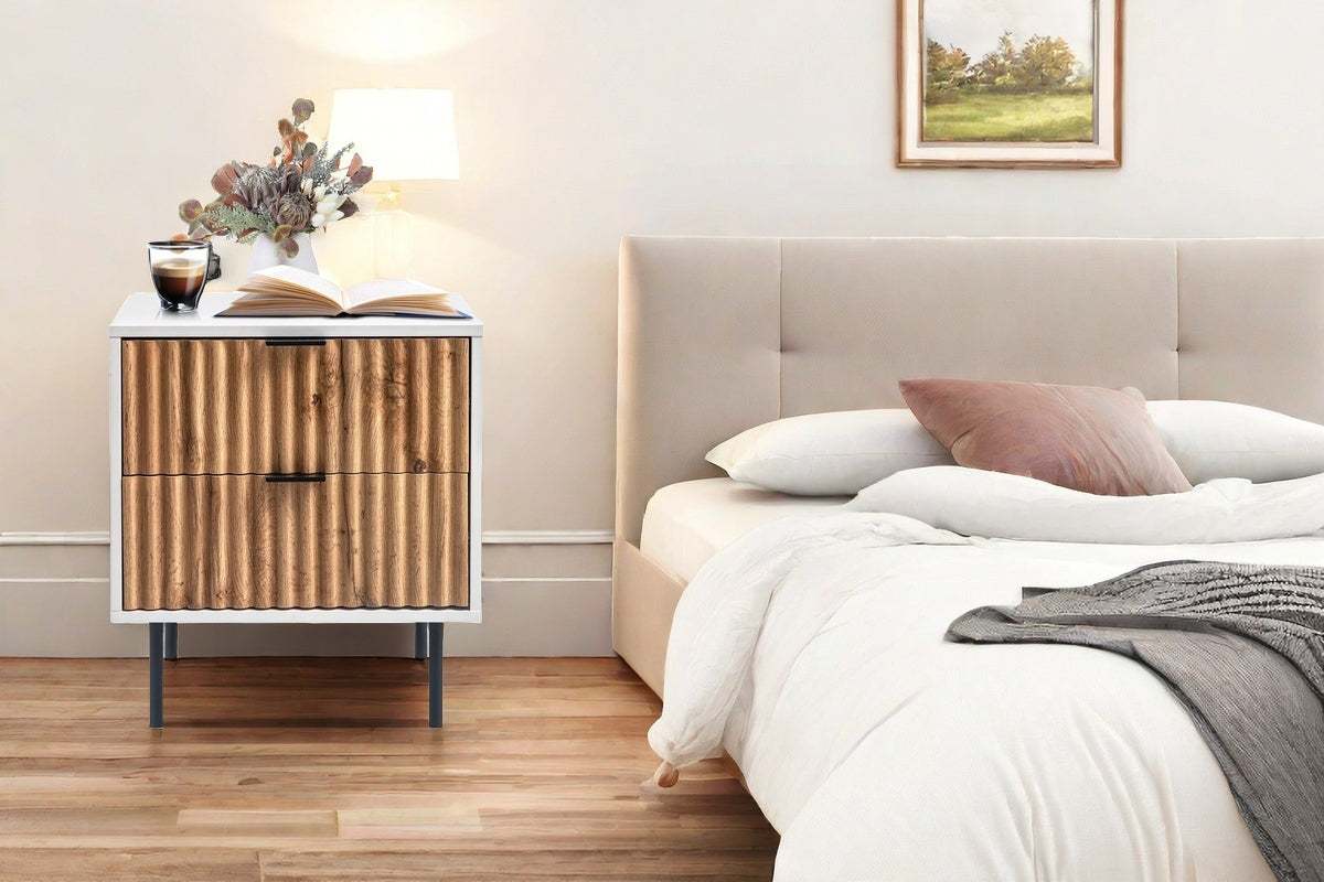 Chelsea 2-Drawer Bedside Table with white and wavy timber facade, sleek black handles, and metal legs next to a bed.