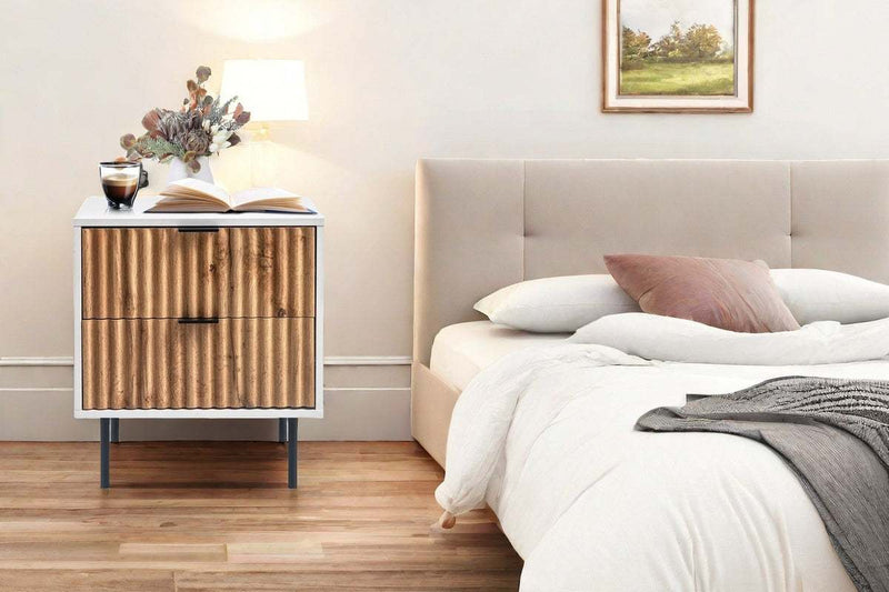 Chelsea 2-Drawer Bedside Table with white and wavy timber facade, sleek black handles, and metal legs next to a bed.